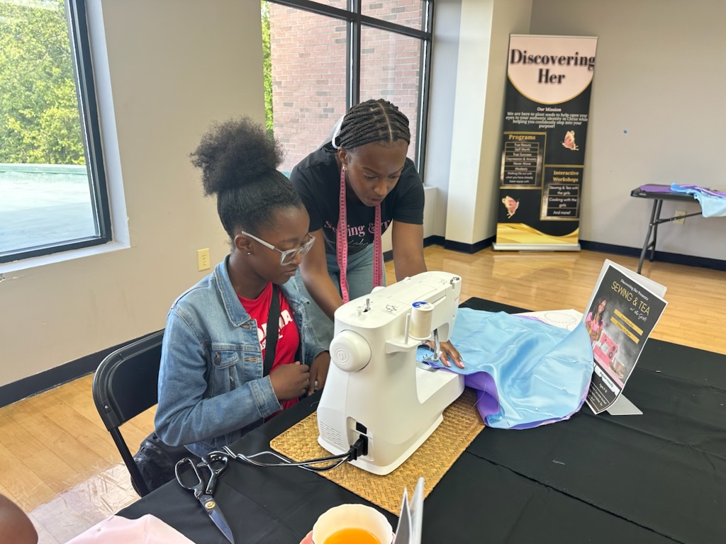 Mia encouraging a girl as she works at a sewing machine